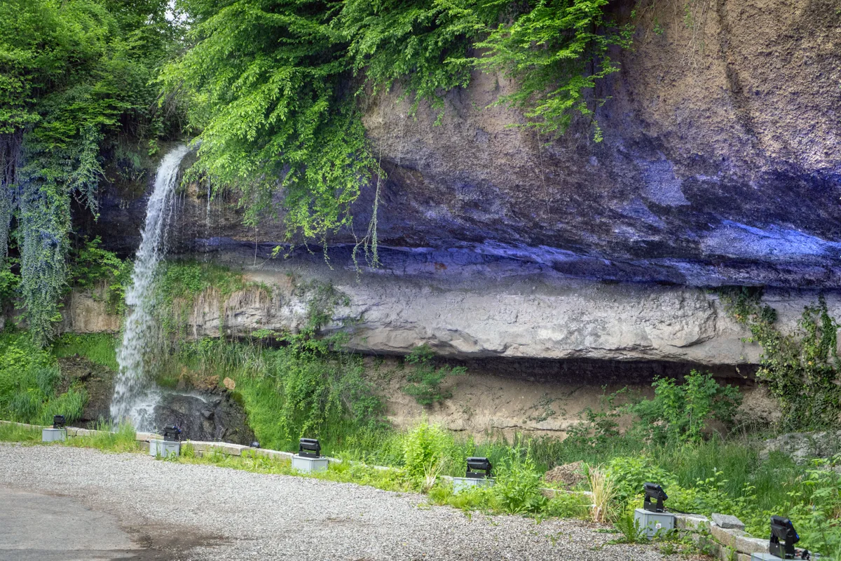 Ein Wasserfall ergiesst sich in einen Weiher. Die Felswand hinter dem Wasserfall wird mit Scheinwerfern blau angestrahlt.